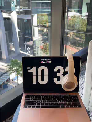 A rose gold laptop sits open on a desk next to a large window overlooking a city. White headphones hang over the laptop screen, which displays a digital clock and the words 'uni diaries'. The scene is bathed in natural light.