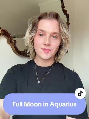 A selfie-style shot of a person with light brown wavy hair, black t-shirt, and a silver necklace. The background consists of a mirror with an ornate gold frame and a wall. Text overlay at the bottom, in a rounded rectangle, reads 'Full Moon in Aquarius'.