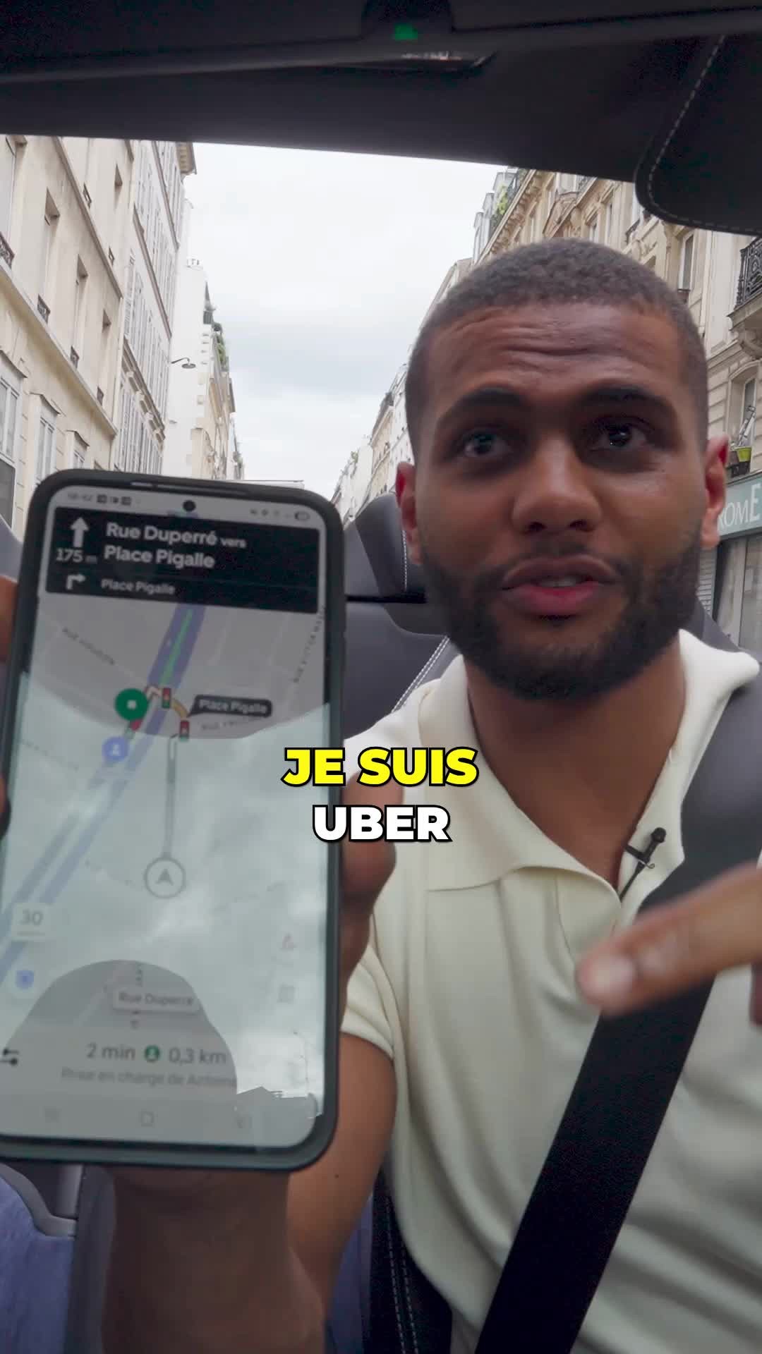 A man in a light-colored polo shirt is holding a smartphone angled towards the camera. The phone screen displays a map with route directions. The background shows a city street with buildings and a car's interior. The text overlay "JE SUIS UBER" is in a white rectangular box in the lower-center of the frame.