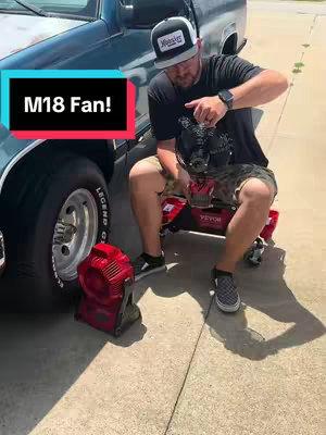 The image shows a man sitting on a red rolling stool next to a truck. There is a red fan on the ground. The man is wearing a hat and black t-shirt and holding a black object with a similar looking object. The background is concrete and grass.