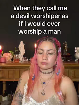 The cover image presents a medium shot of a person with pink hair, heavily accessorized with jewelry and makeup. The background is blurred, suggesting a room with candles and religious statues. Text overlayed on the top says: "When they call me a devil worshiper as if I would ever worship a man".