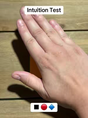 The image shows a hand with the palm facing upward as it covers a small orange block. Above the hand, text reads: Intuition Test. At the bottom of the image, three different colored shapes are visible. The background appears to be a wooden surface.