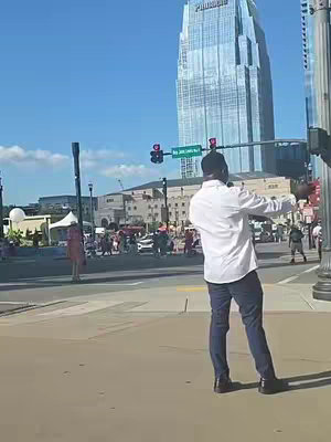A man in a white shirt and blue jeans faces away from the camera, extending his arms out to the sides. He stands on a sidewalk or crosswalk. In the background, a large, multi-story building with a reflective glass facade dominates the scene. The sky is blue with few clouds, and other people are visible. The lighting suggests a sunny day.