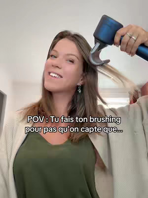 A woman is holding a hair dryer in her right hand, seemingly styling her hair. She is smiling, and the scene is in a well-lit indoor setting with a neutral background.