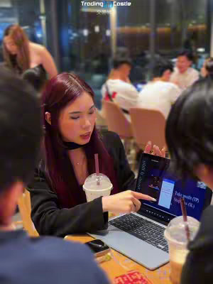A woman with dark red hair is speaking while pointing to a laptop screen, inside a cafe setting. She is holding a drink with a straw. The laptop screen highlights a trading interface.