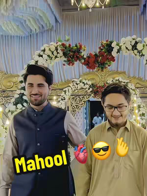 Two men are posing for a photo at a wedding. The man on the left is wearing a navy vest over a cream collared shirt, and the man on the right is wearing a gold shirt and glasses. They are smiling at the camera. There are decorative floral arrangements in the background.