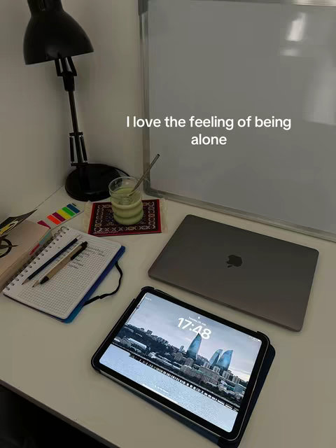 A well-lit, minimalistic workspace with a white desk. On the desk is a laptop, an iPad with a city skyline displayed, a notebook with a pen, a beverage in a glass, and a desk lamp. The color palette is neutral with light tones.