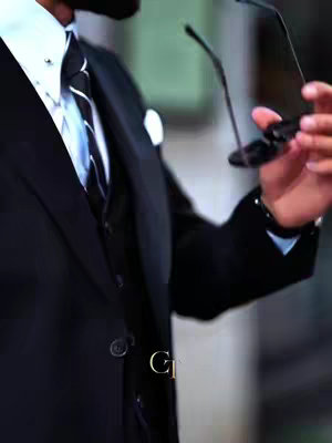 The image shows a person, presumably a man, dressed in a formal suit. The suit is sharply tailored and appears to be black. The person is holding a pair of black sunglasses in their right hand, and the glasses are positioned as if they are about to be put on. The background is out of focus, which puts the focus on the person's attire and the glasses. The overall composition is sophisticated and suggests a stylish or professional setting.