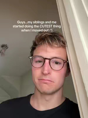 A medium shot shows a young man with fair skin, light brown hair, and glasses looking towards the camera with a slight frown. The background is a light, neutral color, and there's a curtain visible on the right side.