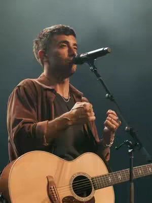 A medium shot depicts a man playing an acoustic guitar on stage, bathed in warm lighting. He is holding the guitar with both hands, positioned in front of him. The man is facing the camera and singing into a microphone. The background is dimly lit, creating a spotlight effect on him and the guitar. The overall color palette is muted, with browns, beige, and darker tones.