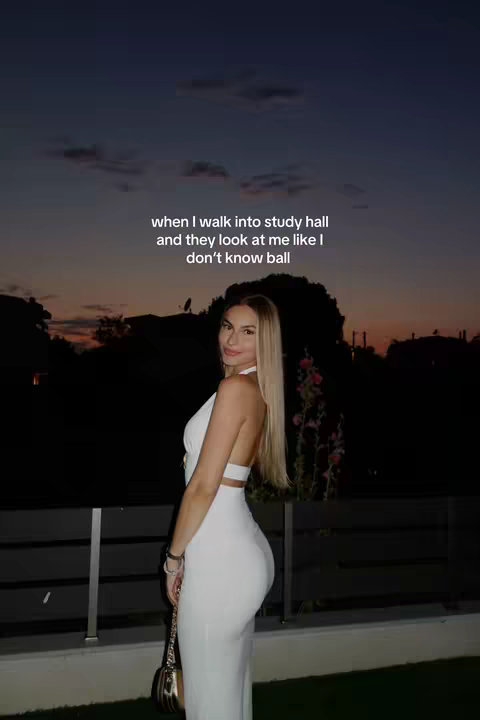 A young woman with long blonde hair is posing outdoors at dusk. She wears a white halter dress, and her long hair flows over her shoulders. The sky is a gradient of orange and purple, reflecting the end of the day. She is positioned slightly off-center, with her head turned towards the camera. A silver handbag hangs from her arm The image has a soft focus and utilizes natural lighting, giving it an aesthetic appeal.