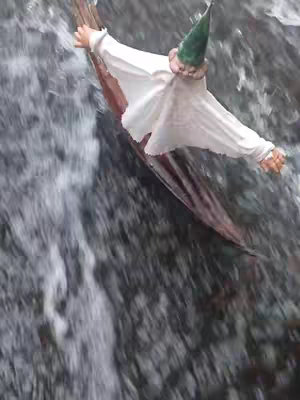 An image from an overhead perspective presents a figure, possibly a gnome or a stylized character, navigating a rapidly flowing stream or waterfall. The figure is positioned as if riding a makeshift raft, crafted from what appears to be a log or a piece of wood. The background is a blur of rushing water, with the grayscale tones emphasizing the movement and the potential danger or excitement of the situation. The character wears a light-colored garment and a pointed green hat. The image is dynamically composed, lending an impression of action and the possibility of adventure or humor.