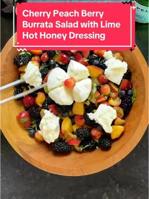 The image is a top-down shot featuring a vibrant salad in a wooden bowl. The salad's focal point includes white burrata cheese, chunks of orange peaches or similar fruits, red cherries, blueberries, blackberries among other fruits and basil leaves. A vibrant red banner with white text displaying the title of the dish is in the image.  The composition utilizes natural lighting, and the dark background adds depth and contrasts with the colourful salad.