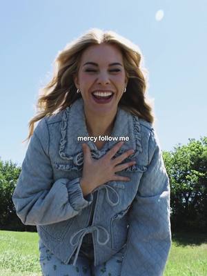 A woman with long blonde hair is smiling widely, her hand placed on her chest. She wears a denim jacket, with a blue sky and green background.