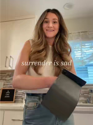 A medium shot features a woman standing in a kitchen setting. She has long, wavy brown hair and is smiling slightly while looking towards the camera. She wears a white sleeveless top and is holding a gray item in her hands. Behind her, a kitchen counter and cabinets are visible, with a small chalkboard sign to her left. Text overlayed across the image reads "surrender is sad".