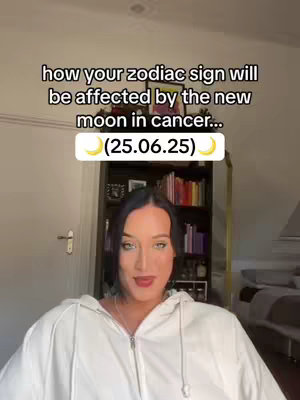 A woman with dark hair and a white hoodie is centered in the frame, smiling gently. Above her, overlaid text in white font discusses astrology and the new moon. A bookshelf filled with books is visible in the background.