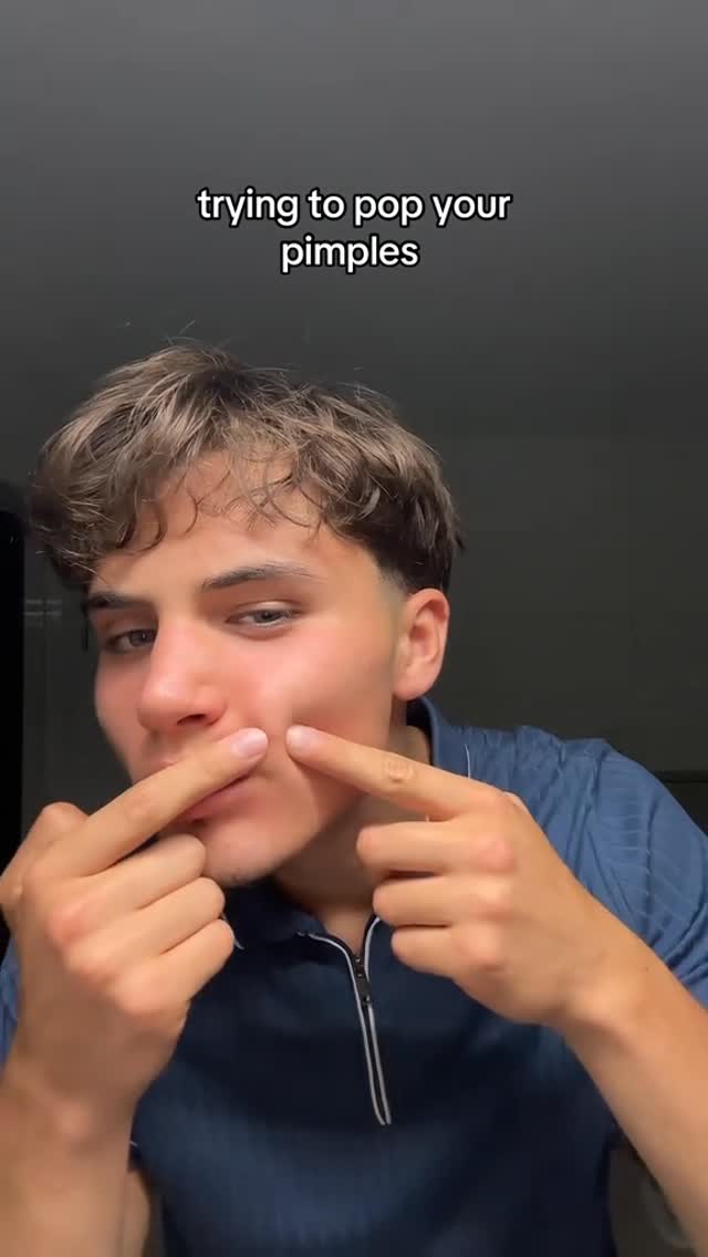 A young man is shown with his face close up against a dark background. The man has light brown hair and is wearing a dark blue collared shirt. His fingers are touching his cheeks, as if he's about to pop a pimple. Text is overlaid on the image, indicating the action depicted in the video.