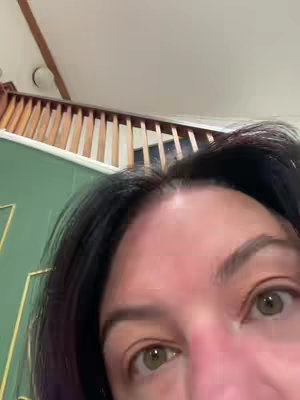 A close-up shot focuses on the upper face of a woman. The background includes a staircase and a glimpse of a green wall. The woman's face fills much of the frame.