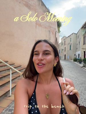 A selfie-style image featuring a woman smiling in an outdoor setting, likely in a European town. She is positioned slightly off-center, with a tan building and a small street in the background. She has long, dark hair and is wearing a black and white polka-dotted top, and gold jewelry. Text overlay is present in the image.