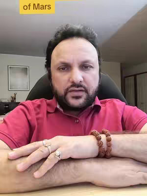 A man with dark hair and a beard is seated, hands resting on a table, wearing a pink shirt and multiple rings, with a beaded bracelet prominent on his wrist. The background includes a framed picture and blurry interior elements.