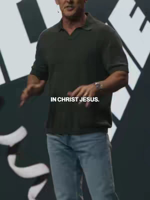 A man stands in front of a blurry background, gesturing. He is wearing a dark green polo shirt and light blue jeans. Text overlayed at the bottom.