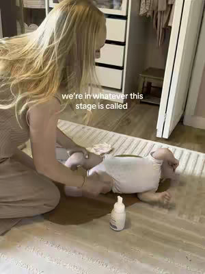 A woman is kneeling on a carpet, attending to a baby lying on the floor. The setting appears to be a bedroom or a closet, with a cupboard in the background. Text overlayed on the image reads "we're in whatever this stage is called".
