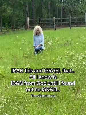 A woman with blonde curly hair kneels in a vibrant green field dotted with small white flowers. She wears a light blue button-down shirt and jeans. Behind her is a wooden fence and a treeline with green foliage under an overcast sky. Text overlays her and the image, with the caption: 'IRAN this and ISRAEL that.. All I know is IRAN from God until I found out he ISRAEL. SherryDamron.org'.