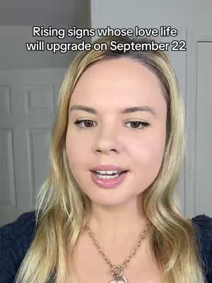 A close-up shot of a woman with blonde hair and fair skin, wearing a dark blue top and a gold necklace with a heart pendant. She is centered in the frame, looking directly at the camera with a neutral expression. The background is a plain white wall with a slightly visible white door frame on the left. The overall composition is simple and focuses on the woman's face.