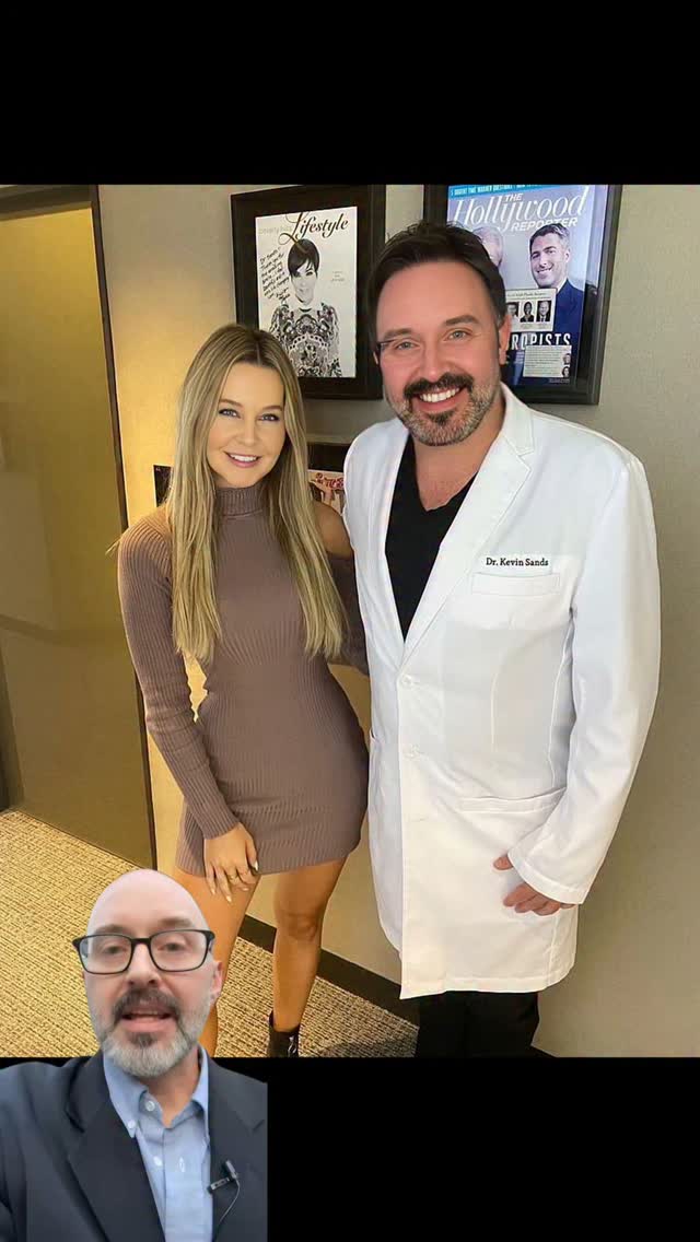 The image features a man and a woman posing together indoors, with a smaller video inset at the bottom showing a close-up of the man's face. The man on the right is wearing a white lab coat and glasses, while the woman on the left has long blonde hair and is wearing a brown dress. The room has a minimalist style with plain walls and a framed magazine cover visible in the background.