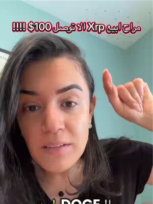 A woman with dark hair and a black top is in a selfie-style video, pointing upwards with her right index finger. The background is a light teal wall. Text in Arabic script is overlaid above her head.