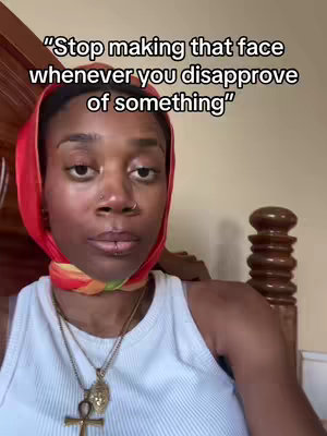 A close-up shot of a Black woman wearing a white tank top and a red headscarf. She looks directly at the camera with a neutral expression, but her gaze suggests a hint of disapproval or weariness. The background includes a hint of a wooden headboard and a wall, with the upper torso and head being the focal point. Overlayed text at the top reads "Stop making that face whenever you disapprove of something".