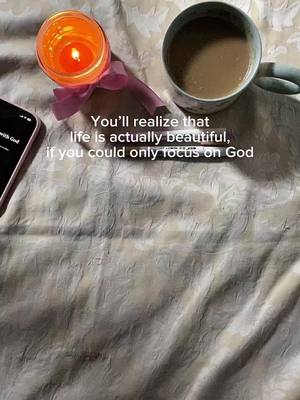 An overhead shot displays a softly lit scene with a white and beige patterned surface. A lit candle in a glass holder with a pink ribbon is to the left, near a partially visible smartphone. To the right, a ceramic mug filled with a light-brown liquid sits. The image's focal point is a message layered over the scene in a handwritten-style serif font.