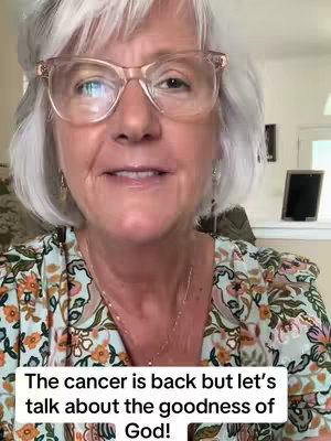 A woman with short white hair and glasses is centered in the frame, smiling slightly. The background is a blurred interior with glimpse of a table, a framed picture and an open door, suggesting a domestic setting. The woman wears a floral patterned top and a delicate necklace. At the bottom of the image, overlaid text provides context.