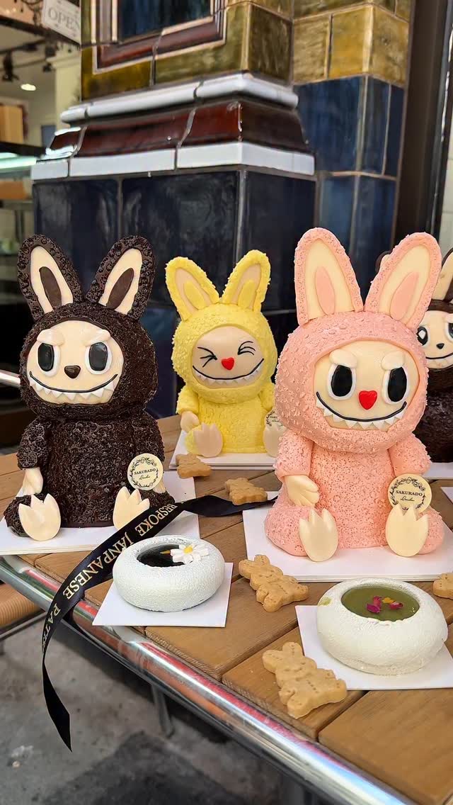 The image showcases a display of Labubu-style cakes in a cafe setting. Three cakes, each designed to resemble the Labubu bunny character, are prominently featured on a wooden table. The cakes vary in color: chocolate, yellow, and pink. Each cake has its own unique design inspired by Labubu. Additionally, there are smaller treats: circular white desserts, and bear-shaped cookies. The background suggests a cafe environment with a patterned wall and ambient lighting.