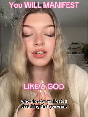 A young woman with long blonde hair is centered in a portrait-style shot. Her eyes are closed, and her hands are clasped together near her chest. Above her head, in vibrant pink text, reads "YOU WILL MANIFEST". Below her, also in pink, is "LIKE A GOD", with the text "who already manifested that thing that you want" in a smaller font overlaid. The background suggests a domestic setting, partially visible with a chandelier and some items on a shelf.