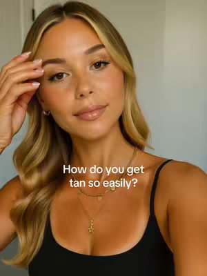 A young woman with blonde hair and fair skin poses for a selfie. She's wearing a black tank top and several gold necklaces. The text reads, 'How do you get tan so easily?' The background is a plain, off-white wall. The warm lighting and the woman's pose suggest a focus on beauty and possibly tanning.
