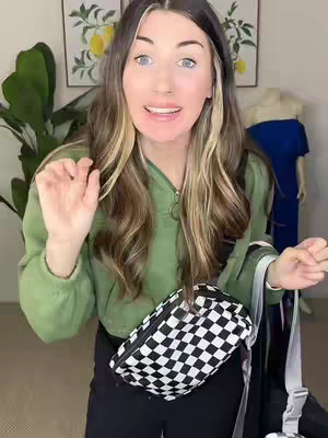 A woman with long hair in a green hoodie is holding a black and white checkered bag in front of her waist, with another black bag in her hand. Behind her are a pair of lemon art pieces and a mannequin with a blue dress.