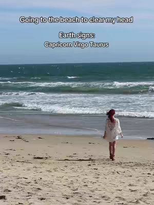 A woman walks away from the camera on a sandy beach. The ocean is in the background with a bright sky.