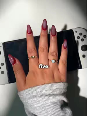 A close-up shot of a hand with perfectly manicured nails. The nails are long and oval-shaped, with a red tip and pinkish-clear base. The hand has three golden rings with gemstones and is positioned over a black and white Nintendo Switch console. An additional text layer 'five' is added between the fingers.