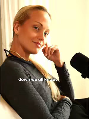 A woman with blonde hair is seated, angled towards the camera. She's wearing a dark top and has a necklace. A microphone is visible to her right. The background appears to be a plain, light color. Text overlayed at the bottom reads 'down we all know.' 
