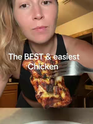 A woman holds a piece of cooked chicken on a fork, presenting it towards the camera. She's looking directly at the viewer, and the chicken is centered in the frame. Text overlayed on the image says, "The BEST & easiest Chicken". The background is slightly blurred.
