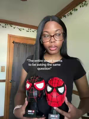 A woman with dark hair and glasses holds two Lego Spider-Man busts, with the Miles Morales and Peter Parker designs. The background shows a room with a window, a door, and some decorative vines.