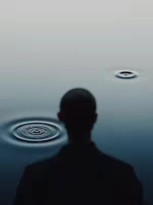 A silhouette of a person looking out over water. The water has several sets of concentric ripples, indicating something has been dropped into the water. The sky is a pale, muted color.