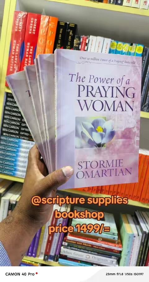 The image showcases a stack of books held in a hand, with a focus on the top book titled 'The Power of a Praying Woman' by Stormie Omartian. The cover art features a close-up of a purple flower. The background is a bookshelf filled with various books. The hand holding the books is visible, and there is added text below the books that says '@scripture supplies bookshop price 1499/='.