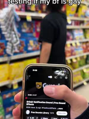 The image shows a smartphone displaying the Grindr app in the foreground, held by a hand, with a person standing in front of shelves in a store in the background. The phone screen features the Grindr logo and a play button for a video related to a Grindr notification sound. Above the phone, text reads 'testing if my bf is gay'. The background is a supermarket aisle with various products on shelves.