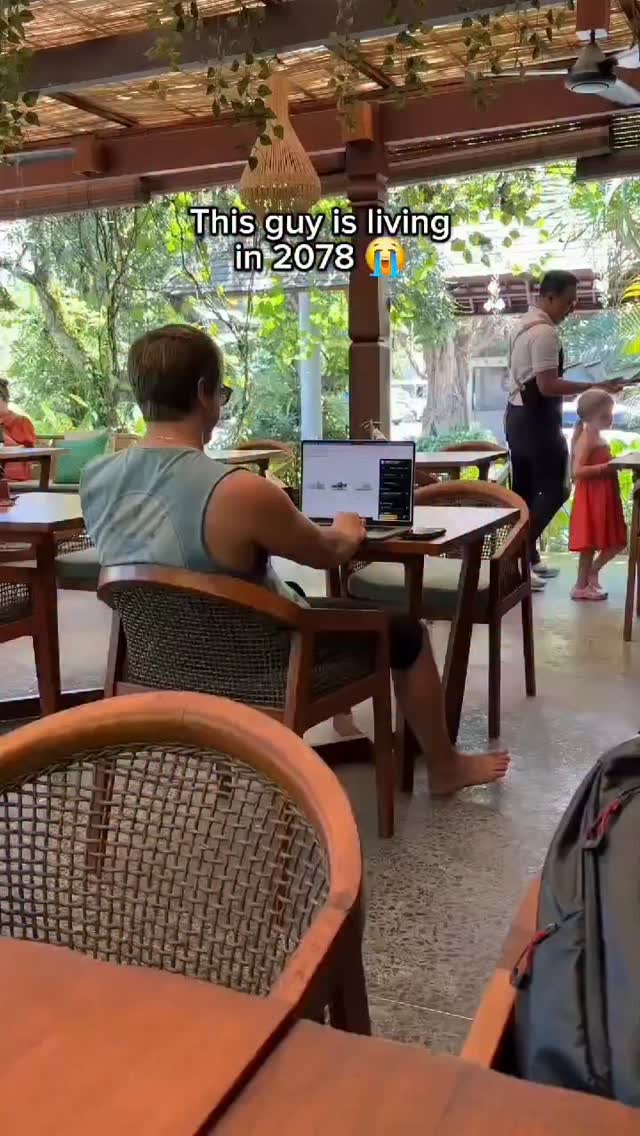 A man is sitting at a table outdoors, using a laptop. The scene is framed from behind the man, showing his bare feet. Text overlayed reads "This guy is living in 2078." A waiter and a young girl in a red dress stand in the background. The immediate setting appears to be a cafe or restaurant, with tables, chairs, and overhead cover. The background includes lush greenery.
