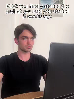 A young man with dark hair and a black t-shirt sits in front of a computer, his face displaying a shocked and stressed expression. He appears to be looking at lines of code or a spreadsheet, implying work-related stress or a project deadline.