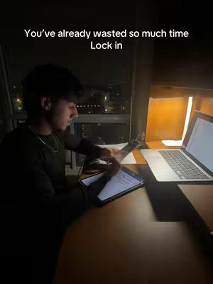A young man is studying at a desk, illuminated by a desk lamp and the glow of a laptop screen. He is focused on some papers, suggesting he is working on a project or studying. The background is blurry and dark, but city lights are visible through a window.