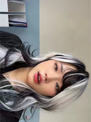 A selfie-style shot of a person with striking black and white hair, angled slightly to the left. They have fair skin, full lips with pink lipstick, and are wearing some basic makeup. The background shows a light gray wall and off-white closet or shelf.