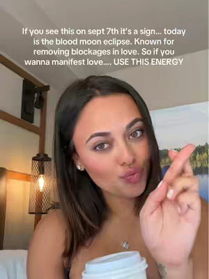 A woman with long, dark hair and light skin is in a selfie-style shot. She holds a coffee cup in her left hand, close to her chest. Her right hand has fingers crossed, and she has a slight pout. The background includes a room with a painting, a lamp, and a bed.